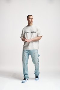 Beige ylisuuri t-paita, jossa musta "2Y STUDIOS" -painatus, vaaleansiniset cargo-farkut ja mustat sekä vaaleansiniset lenkkarit, tasaisen taustan edessä.