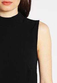 Svart ärmlös topp med hög krage, tillverkad av ett mjukt och lätt material. Har en figurnära silhuett och rena kanter.