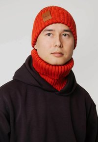 Czerwona dziana czapka z prążkowaną fakturą i brązową skórzaną naszywką. Dopasowany prążkowany czerwony komin na szyję. Noszona na czarnej bluzie z kapturem.