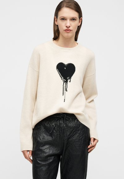 Γυναίκα που φοράει εκρού πουλόβερ με μαύρη γραφική καρδιά που στάζει, συνδυασμένο με μαύρο υφασμάτινο παντελόνι με κορδόνι, πάνω σε απλό φόντο.