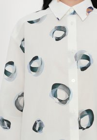 Bílá košile s knoflíky, která má abstraktní kulaté vzory v odstínech modré a zelené. Lehká, poloprůsvitná tkanina s klasickým designem límce.
