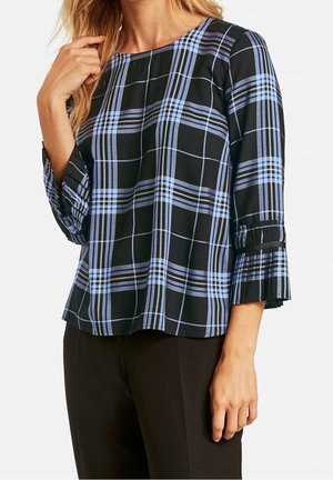 Femme portant un chemisier à manches longues à carreaux noirs et bleus avec des détails plissés sur les manches, associé à un pantalon noir.