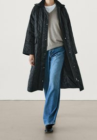 Zwarte gewatteerde lange jas met drukknopen, gedragen over een beige v-hals trui, gecombineerd met lichtblauwe wijde jeans en zwarte schoenen.