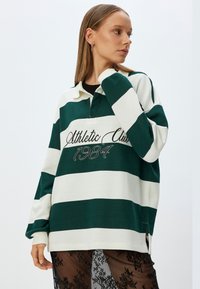 Vihreä ja valkoinen raidallinen collegepaita, jossa on kaulus ja etuaukko. Paidan etuosassa on "Athletic Club 1984" -brodeeraus. Pehmeä kangas, oversized-mallia.
