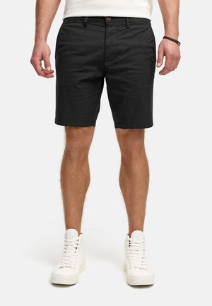 Schwarze Shorts aus leichtem Material mit geradem Schnitt. Mit einem knöpfbaren Bund und Seitentaschen. Getragen mit weißen High-Top-Sneakern.