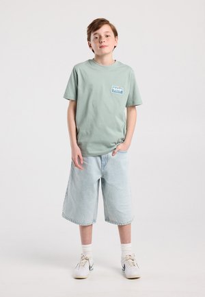Jongen staat met één hand in de zak, draagt een mintgroen T-shirt met de tekst "Surf", lichtblauwe denim shorts tot op de knie, witte sokken en sneakers.