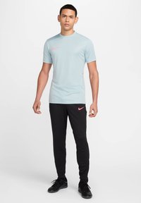 Šviesiai mėlyna sportinė marškinė su rožiniu Nike logotipu, derinama su juodomis siaurinančiomis sportinėmis kelnėmis su rožiniu akcento logotipu, pagaminta iš glotnaus, lengvo audinio.
