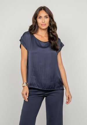 Femme aux longs cheveux bruns portant une blouse ample bleu marine et un pantalon assorti, debout devant un fond uni gris clair.