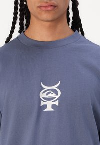 Blå bomulls t-skjorte med rund hals, med et stort hvitt grafisk logo-design på brystet og en jevn tekstur.