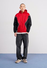 Rød og sort vindjakke med hætte, med hvide detaljer, parret med mørke jeans og brune sneakers på et blåt gulv.