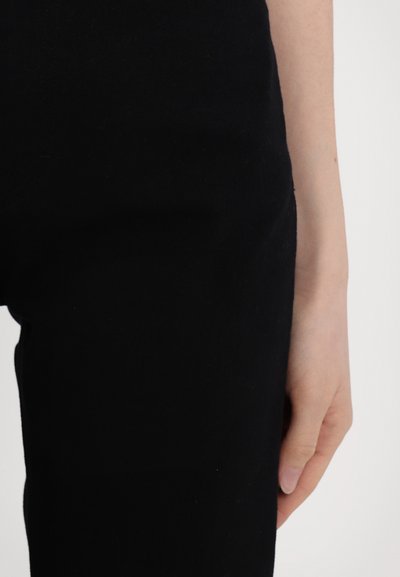 Sorte denimbukser med glatt tekstur, tettsittende design og minimale sømdetaljer, med en nærbilde av midjebåndet og hånden.
