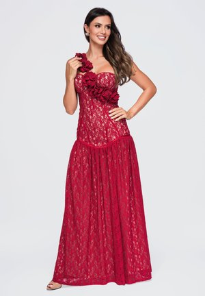 Femme portant une robe rouge en dentelle sans bretelles avec des applications florales sur le corsage, debout avec une main sur la hanche et souriante.