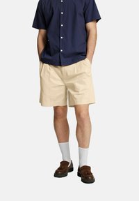 Pantaloni corti beige in cotone con pieghe, tasche laterali, abbinati a una camicia blu navy a maniche corte. Indossati con calze bianche e mocassini marroni.