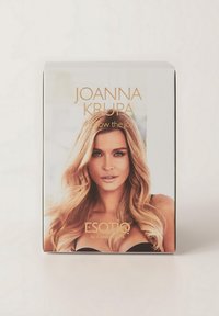 Pudełko produktu z "Joanną Krupą" w złotym napisaniu. Duży obraz kobiety z długimi, falowanymi blond włosami. Tło jest rozmyte.