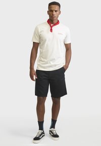Homme portant un t-shirt blanc avec un texte rouge, un short noir, des chaussettes noires avec des flammes bleues, des baskets noires et blanches, et un foulard rouge autour du cou.