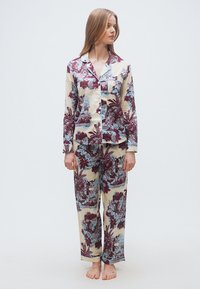 Γυναίκα που στέκεται ξυπόλητη φορώντας πυτζάμα με μακρυά μανίκια και κουμπιά, σετ με μπλε και βουργουνδί ανθικά και τοπία μοτίβα σε εκρού φόντο.