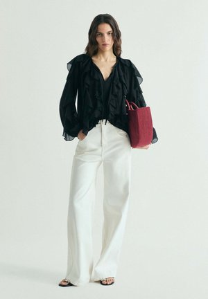 Mujer con blusa negra con volantes y pantalones anchos blancos, sosteniendo un bolso tote texturizado de color rojo oscuro, de pie frente a un fondo liso y claro.