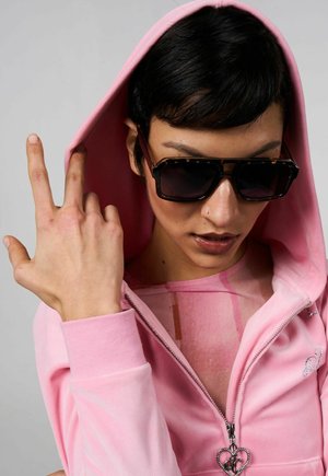 Felpa rosa in velluto con zip e un grande cappuccio, dotata di un tirante a forma di cuore. Indossata sopra un top trasparente, accessorata con occhiali da sole scuri.