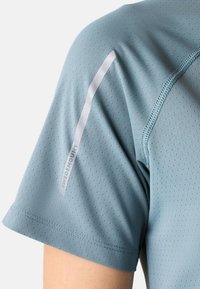 Lichtblauw sportshirt met geperforeerde textuur, korte mouwen en reflecterende accenten langs de schouder. Bevat subtiele merkmarkering.
