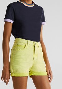 Personne portant un t-shirt bleu marine avec un col bordé d’arc-en-ciel et un short en jean vert clair à ourlet retroussé, debout devant un fond uni.