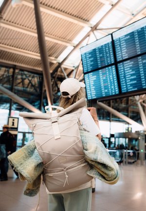 Eine Person mit einem beigen Rucksack und einer Kappe schaut sich die Abflugtafeln in einem modernen Terminal mit Glaswänden und Metallträgern an.
