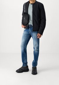 Musta bomber-takki, vaaleansininen collegepaita, kulutetut siniset farkut ja mustat tennarit. Oikeassa kädessä logoa kantava laukku.