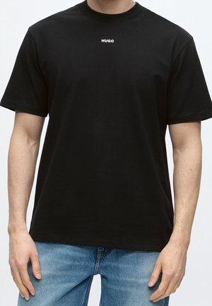 Mand iført en almindelig sort t-shirt med korte ærmer med teksten "HUGO" centralt ved halsudskæringen og lyseblå jeans.