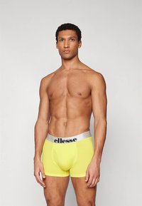 Mišićav muškarac stoji, odjeven u svijetlo žute boksačke gaće sa srebrnom pojasevima i "ellesse" logotipom, na jednoličnom svijetlom pozadini.