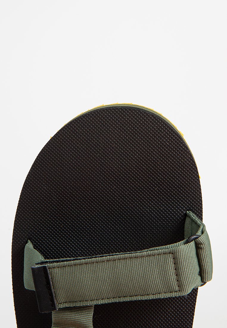 Zielone i czarne sandały z teksturowaną gumową podeszwą, regulowanym paskiem i zaokrąglonym noskiem. Wyraźny żółty akcent wzdłuż krawędzi.