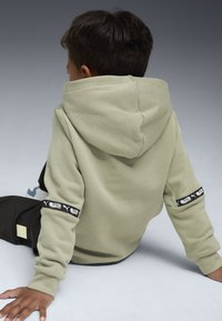 Bambino seduto per terra con la schiena rivolta verso la telecamera, che indossa un felpa verde chiaro con toppe nere sulle maniche e pantaloni neri.