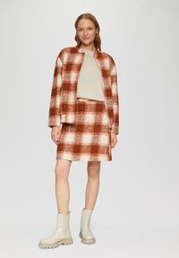 Karo-Jacke und Rock-Set in Rost- und Creme-Tönen. Die Jacke hat einen Stehkragen; kombiniert mit hellbeigen Stiefeln mit klobigen Sohlen.