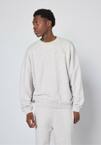 Svetlo siva majica s dugim rukavima, klasičnog izreza, slobodnog kroja, sa rebrastim manžetama i donjim rubom, koja sadrži suptilan logo. Model nosi odgovarajuće dukserice ispod.