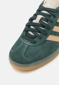 Zelené semišové tenisky s tmavě zelenými tkaničkami, béžovými kožen�ými pruhy a světle hnědou gumovou podrážkou. Obsahují prošívané detaily a štítek s logem.