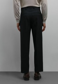 Pantalon noir en tissu texturé, avec une coupe droite et deux poches arrière. Porté avec un pull léger et des chaussures marron.