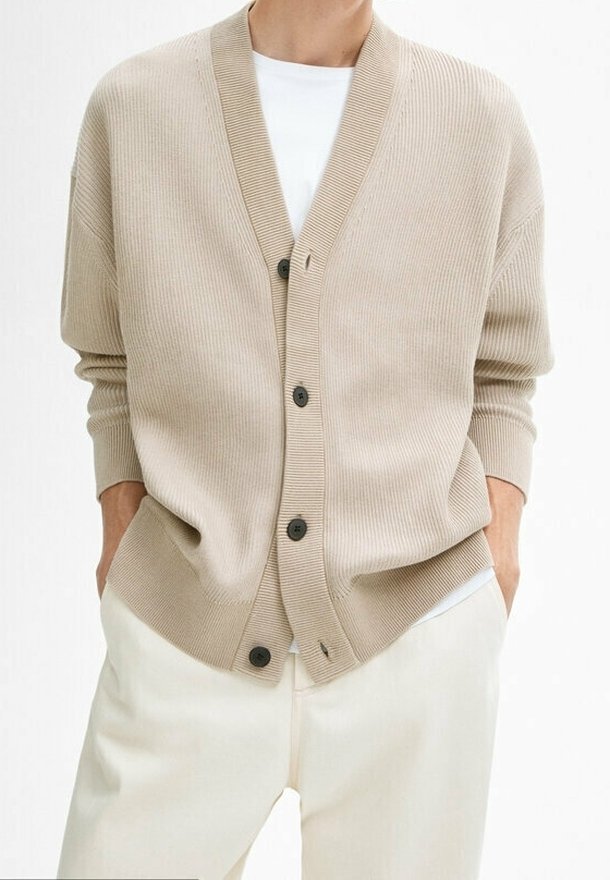 Beige gerippter Cardigan mit V-Ausschnitt, vorderen Knöpfen und lockerer Passform. Getragen über einem weißen T-Shirt mit langen Ärmeln und seitlichen Taschen.