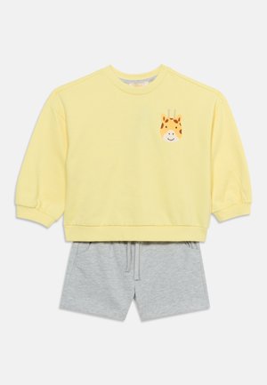 Żółta koszula z długim rękawem z uśmiechniętą twarzą żyrafy na piersi, zestawiona z jasnoszarymi szortami na troczki.