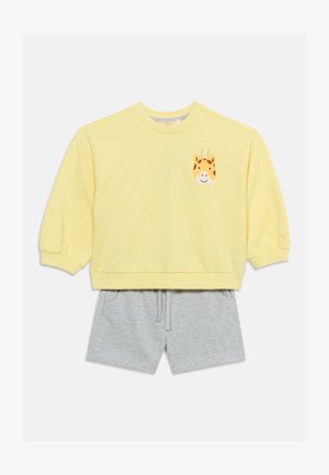 Chemise jaune à manches longues avec un visage de girafe souriante sur la poitrine, associée à un short gris clair à cordon.