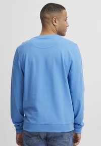 Sudadera azul claro con cuello redondo, manga larga y puños de canalé. Textura suave, ajuste casual. Detalles de costura visibles en la parte posterior.