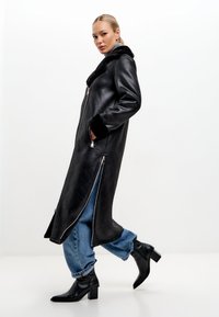 Černý kožený dlouhý kabát se zipem vpředu, kožíškovým límcem a manžetami. Nesený přes volné modré džíny a černé kotníkové boty na podpatku.