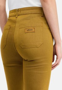 Senfgelbe Jeanshose mit hoher Taille, sichtbaren Nähten und einer Gesäßtasche mit markiertem Etikett. Glatte Textur, figurbetonter Schnitt.