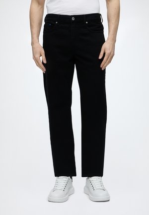 Homme portant un pantalon noir droit et des baskets blanches sur un fond clair uni.