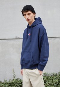 Felpa blu navy in tessuto morbido con tasca frontale e cappuccio con lacci, con un piccolo logo ricamato. Abbinata a pantaloni beige chiaro.