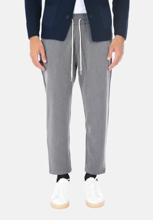 Uomo che indossa pantaloni grigi con coulisse, un maglione blu navy, calze nere e scarpe da ginnastica bianche, in piedi su uno sfondo bianco.