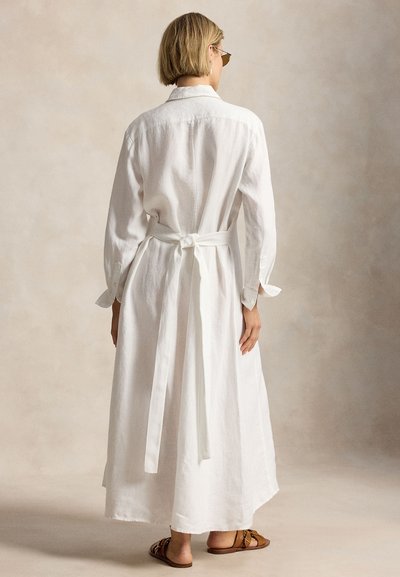 Γυναίκα με κοντά ξανθά μαλλιά, ντυμένη με μακρύ λευκό φόρεμα με ζώνη και καφέ σανδάλια, όρθια με την πλάτη προς το φακό σε ουδέτερο φόντο.