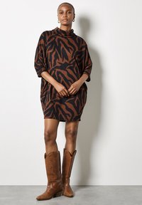 Femme portant une robe courte à imprimé animal noir et marron et des bottes de cowboy en cuir marron jusqu'aux genoux, se tenant contre un mur blanc.