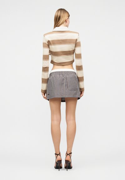 Γυναίκα με καφέ και κρεμ ριγέ cropped μακρυμάνικο πουκάμισο, κοντή καρό φούστα και μαύρες ψηλοτάκουνες σανδάλες, γυρισμένη με την πλάτη σε λευκό φόντο.