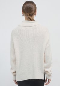 Frau mit braunen Haaren, die zu einem tiefen Dutt gebunden sind, trägt silberne Creolen und einen lockeren, cremefarbenen Strickpullover, wobei sie abgewandt ist.