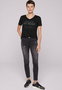 Camiseta negra de cuello en V con el texto "RELAX Ámate a ti mismo", combinada con jeans oscuros ajustados con detalles impresos. Incluye zapatillas negras.