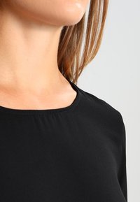 Svart långärmad topp med rundad halsringning, tillverkad av mjukt tyg med en slät textur och subtila sömdetaljer vid nederkanten.