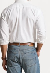 Camicia bianca di cotone con maniche arrotolate, abbinata a jeans in denim blu chiaro e una cintura in pelle marrone, con vista posteriore e dettagli delle tasche.